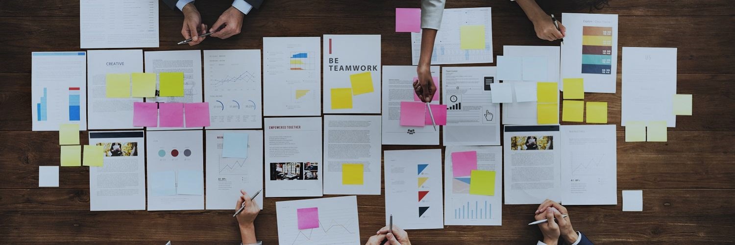 Overhead view of people gathered around a table covered with charts, documents, and colorful sticky notes during a collaborative team meeting.