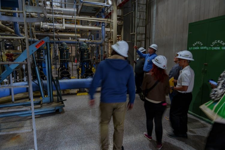 Group of visitors wearing white hard hats stand inside an industrial power plant facility, gathered around a staff member who is gesturing toward large blue and gray pipes, pumps, and valves arranged in a complex network. 