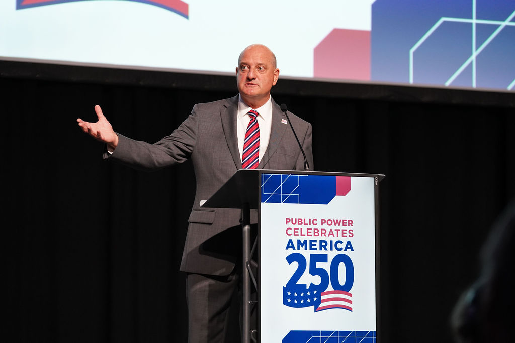 APPA President & CEO Scott Corwin in a suit and red striped tie, standing and gesturing at a podium adorned with a "Public Power Celebrates America 250" sign during his address to the opening general session of APPA's National Conference in Boston, Massachusetts, June 29, 2026. 