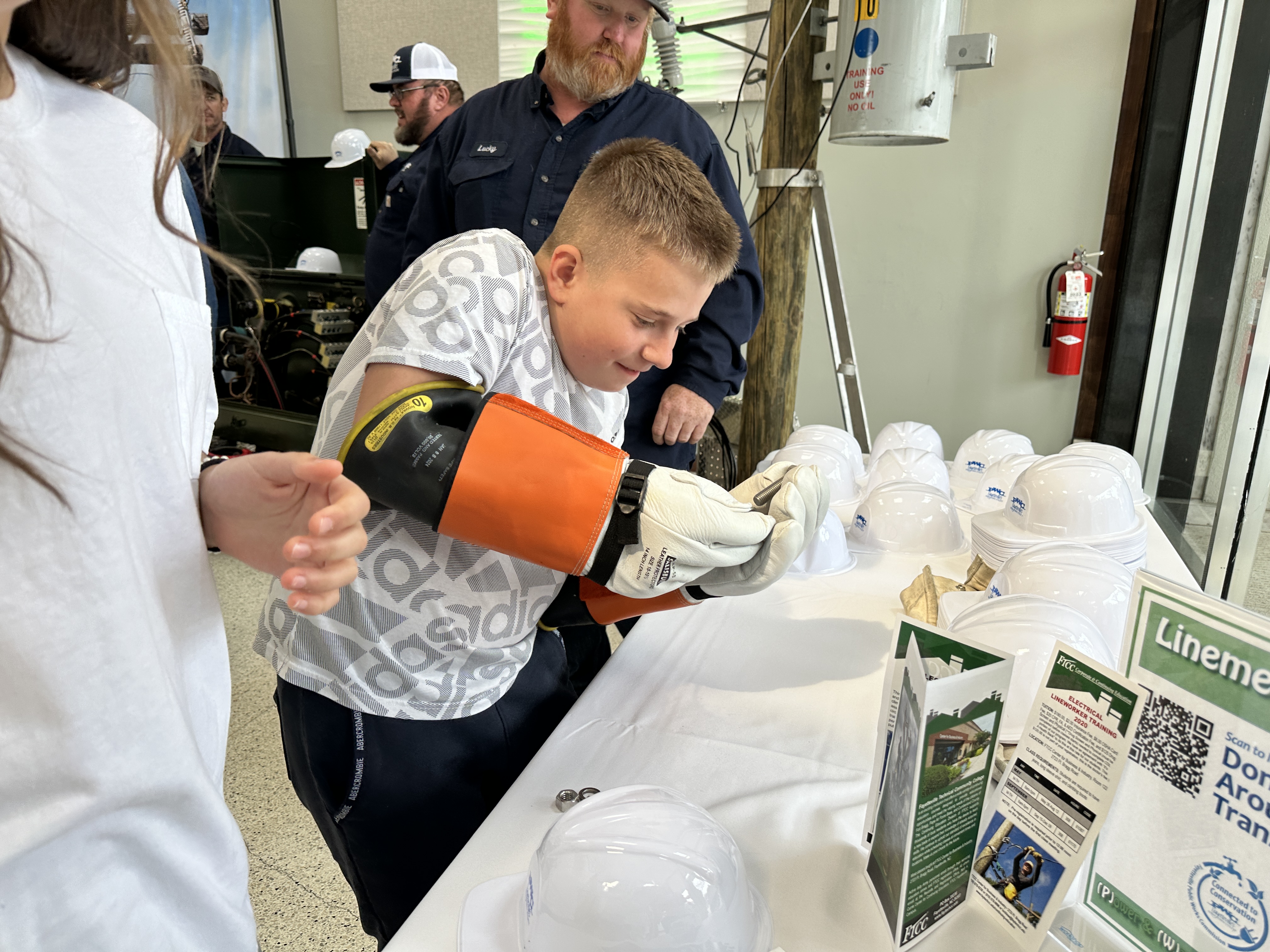 A boy tries on lineworker gear at the PWC Expo while a lineworker looks on.