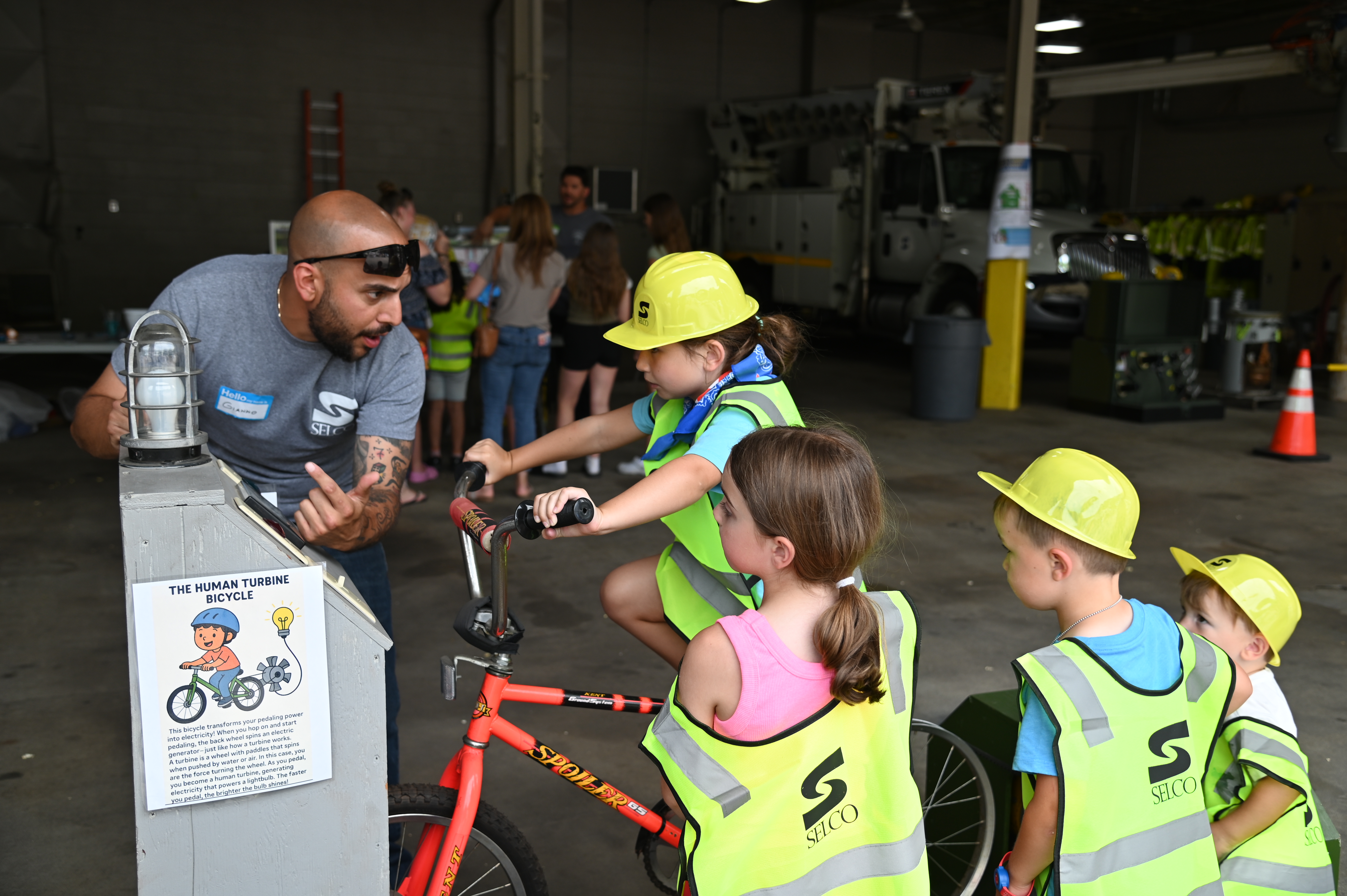 A girl rides a bicycle hooked up to a light bulb while three other children look on, along with an employee of SELCO