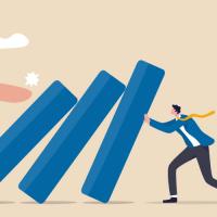 Large hand pushing on row of dominoes while small person tries to hold them up