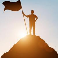 A person stands atop a hill, holding a flag, silhouetted against a low sun in the background. 