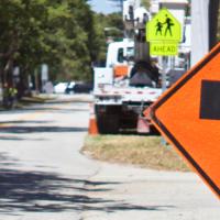 An orange work zone sign of a person holding a flag in front of a road with parked cars