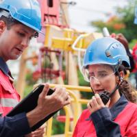 Two people in hardhats looking at a tablet while another worker in safety gear rides a bucket truck in the background