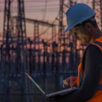 A man in a hard hat and hi-vis vest works on a laptop with distribution lines in the background