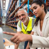With a background of a large warehouse, one person in business attire points to a computer screen as another person in a hi-vis vest watches.