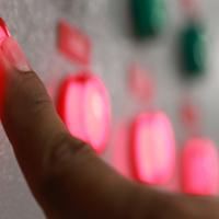 Person pushing a button lit up in red on a control panel filled with red and green buttons