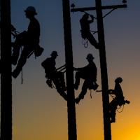 Silhouetted lineworkers atop power poles in front of a sunset