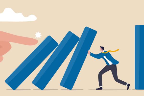 Large hand pushing on row of dominoes while small person tries to hold them up