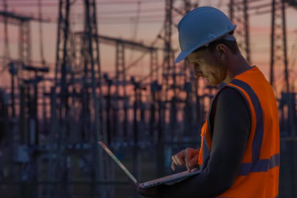 A man in a hard hat and hi-vis vest works on a laptop with distribution lines in the background