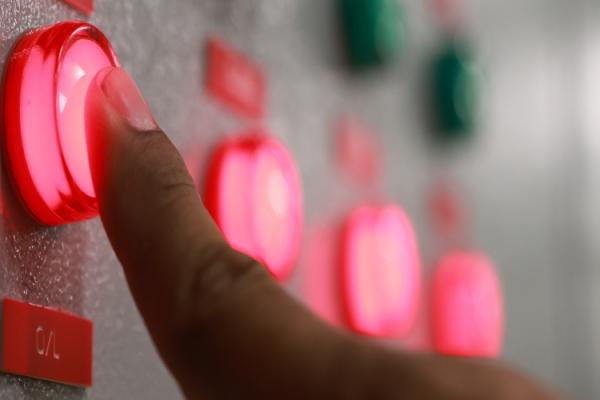 Person pushing a button lit up in red on a control panel filled with red and green buttons