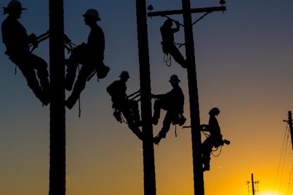 Silhouetted lineworkers atop power poles in front of a sunset
