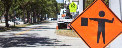 An orange work zone sign of a person holding a flag in front of a road with parked cars