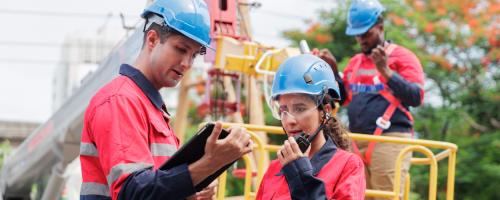 Two people in hardhats looking at a tablet while another worker in safety gear rides a bucket truck in the background