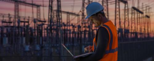 A man in a hard hat and hi-vis vest works on a laptop with distribution lines in the background