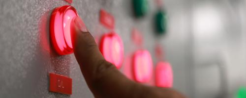 Person pushing a button lit up in red on a control panel filled with red and green buttons