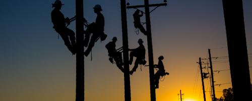 Silhouetted lineworkers atop power poles in front of a sunset